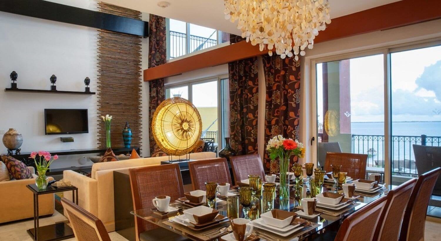 A dining and living area with a rectangular glass dining table set with white and brown dishes, eight dining chairs, a shell chandelier, a living area with a plush sofa and chairs, and flat screen TV, and sliding doors leading out to a patio with furniture and ocean views.
