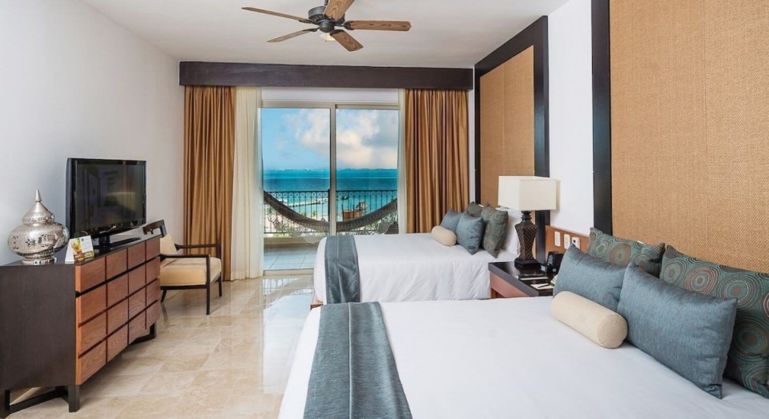 A bedroom with two queen beds with white and blue bedding, a nightstand and lamp in between the beds, a dresser and flat screen TV on the opposite wall, an armchair next to the dresser, and sliding doors leading out to a private terrace with balcony and beautiful turquoise ocean views.