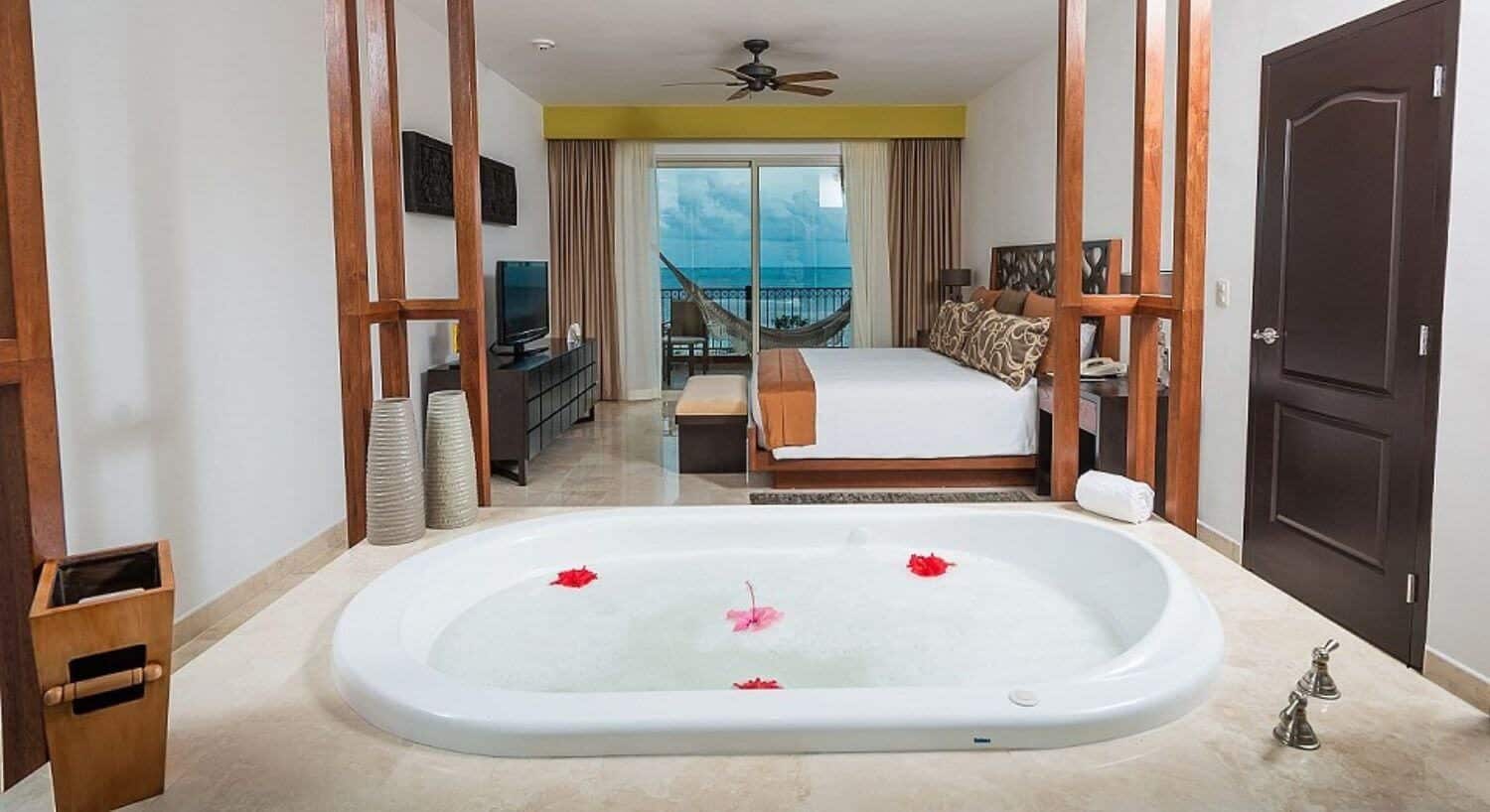 A bedroom with a king bed with white and tan bedding, nightstands and lamps on either side, a plush armchair and lamp in a corner, a jetted tub in the foreground, and sliding glass doors leading out to a balcony with hammock and ocean views.