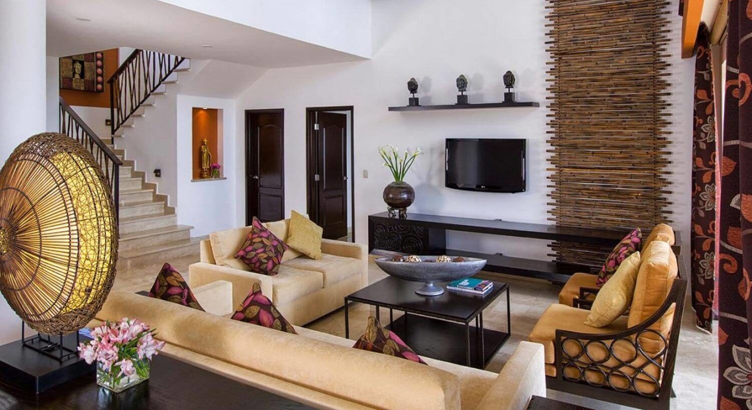 A comfortable living room with tan sofas and chairs, a coffee table and sideboard, a flat screen TV on one wall, and stairs leading up to a second floor.