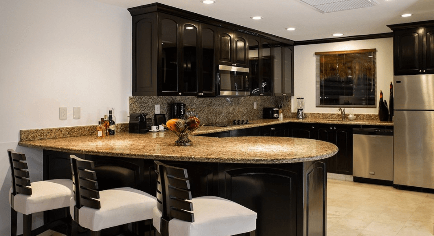 A gourmet kitchen with dark wood cabinets, granite countertops, three white and brown barstools, and stainless steel appliances.