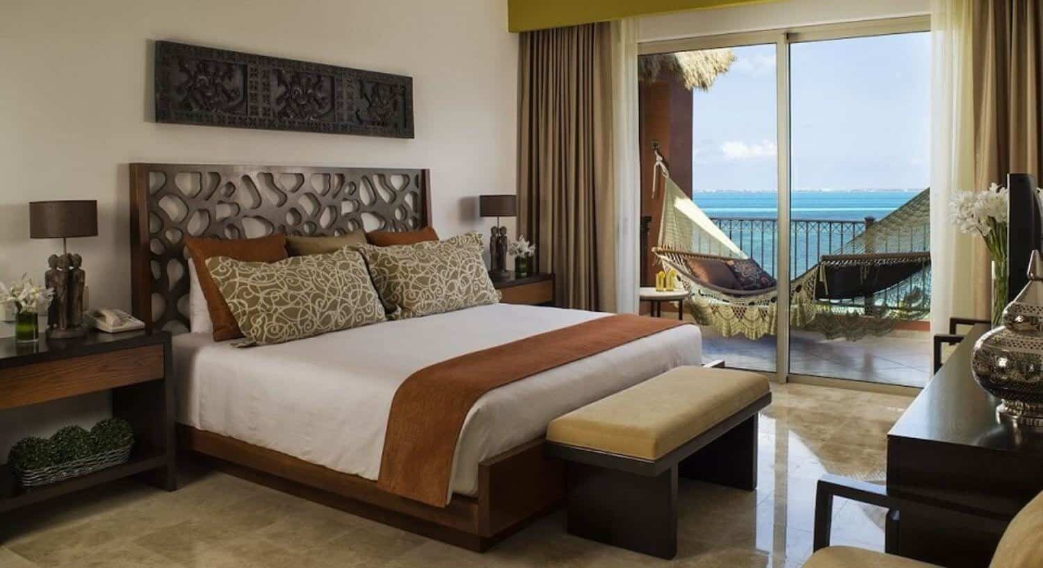 A bedroom with a King bed with white brown and green bedding, a plush sitting bench at the foot of the bed, nightstands and lamps on either side of the bed, a dresser with flat screen TV on the opposite was as the bed, and sliding doors leading out to a balcony with an oversized hammock, and beautiful turquoise ocean views.