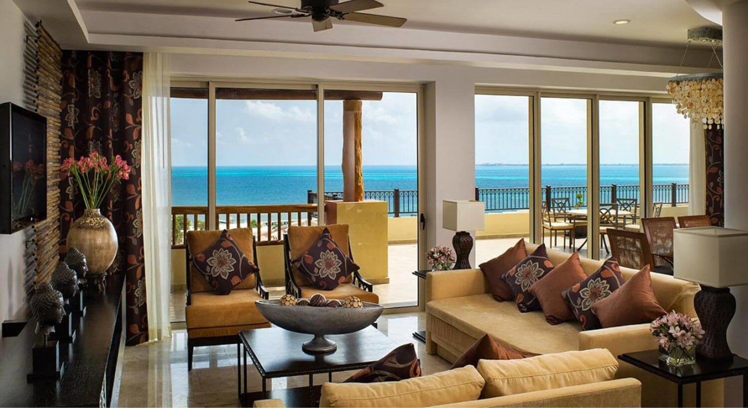 A comfortable living room with plush tan sofas and chairs, a dresser and TV along one wall, a dining area behind the living area, and sliding doors leading out to a large private terrace with patio furniture, and stunning views of the turquoise Caribbean sea.