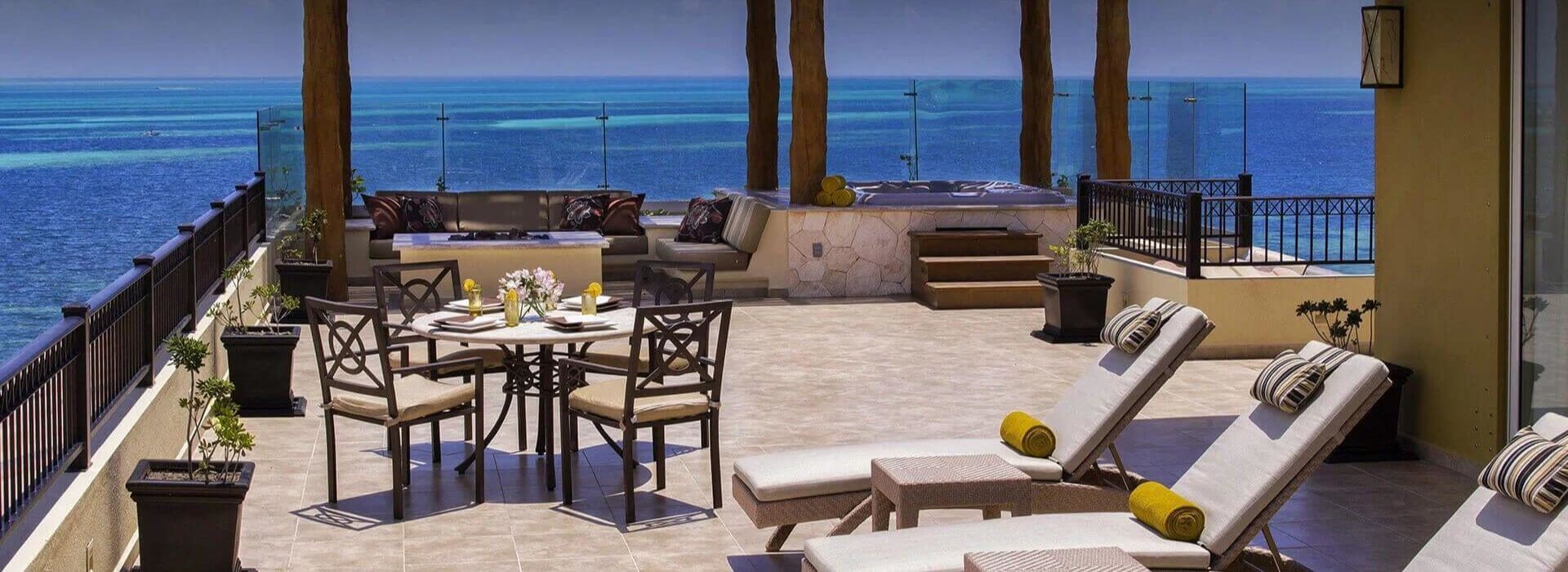 A spacious terrace with lounge chairs, a dining table and chairs, a sitting area with sofas and a fire pit, and a hot tub, and beautiful blue ocean views.