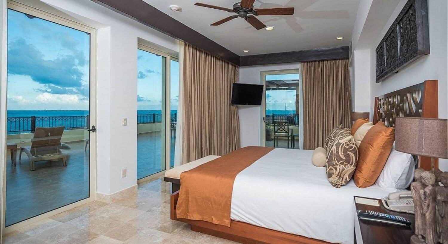 A bedroom with a King bed with white, orange and brown bedding, several sliding glass doors leading out to an expansive wrap around terrace with patio furniture and ocean views.