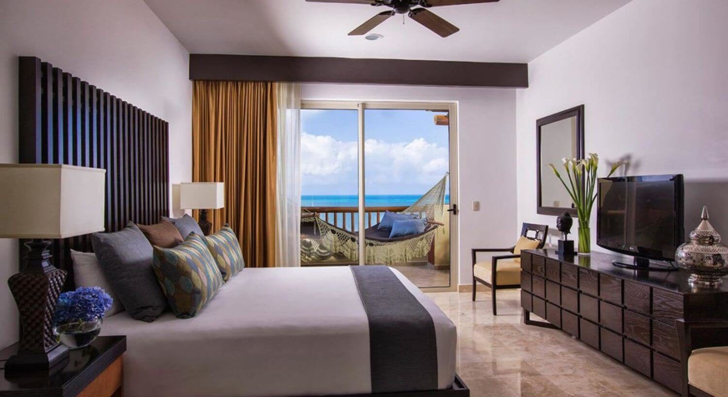 A bedroom with a King bed with white, grey bedding with multi colored throw pillows, nightstands and lamps on either side of the bed, a rich deep wood dresser and flat screen TV on the other side of the bed, and sliding doors leading out to a balcony with an oversized hammock and turquoise ocean views.