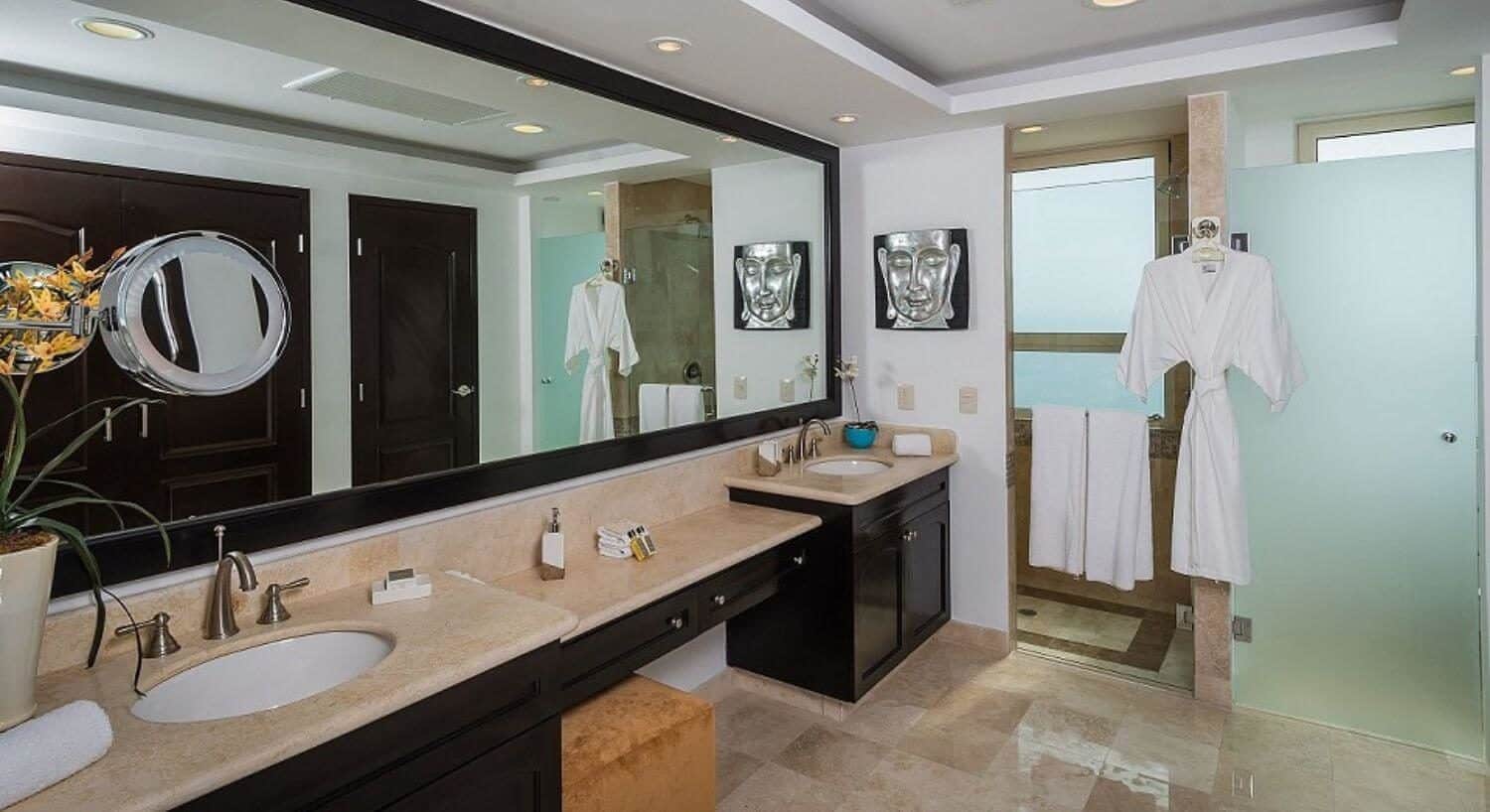 A bathroom with granite countertop with double sinks and a vanity in between them, a full length mirror over the counter, and a walk in shower with a white robe hanging next to the shower.