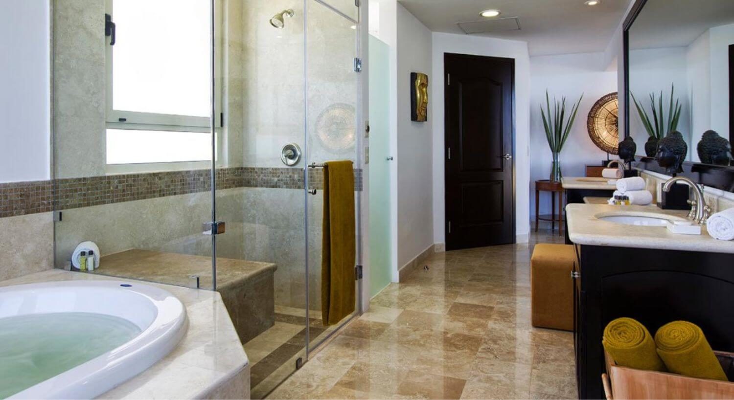 A spacious bathroom with double granite topped sinks, a deep soaking tub, a walk in shower with bench, and rolled gold towels in a basket.