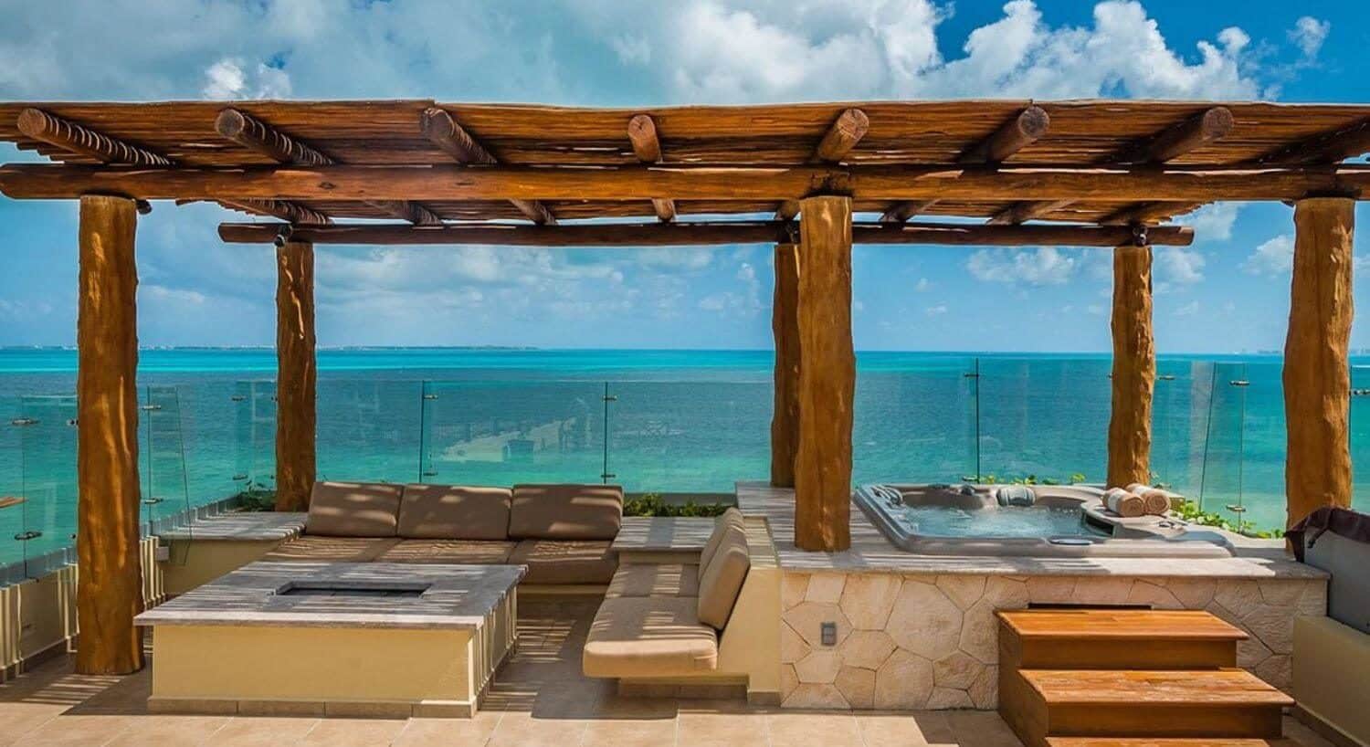 An outdoor living area with couches, a firepit and a hot tub, with a pergola roof and sweeping turquoise ocean views.