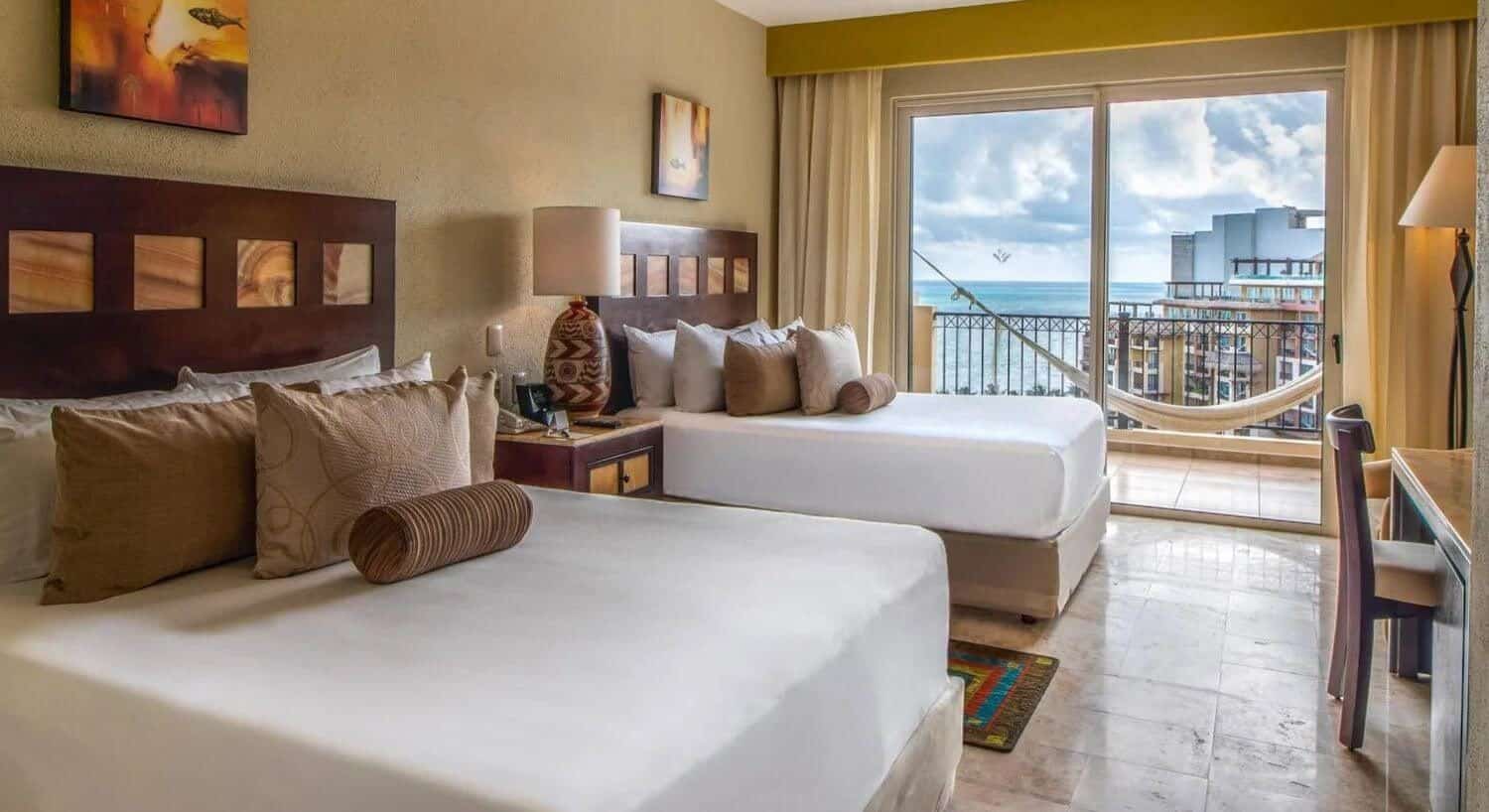 A bedroom with two queen beds with white and brown bedding, a nightsand with lamp in the middle, a writing desk and chair, a plush armchair and floor lamp in the corner, and sliding doors leading out to a private balcony with hammock and resort and ocean views.