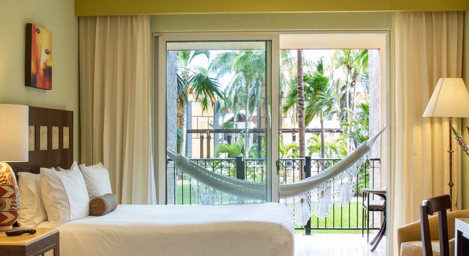 A bedroom with a bed with white bedding, nightstand with lamp, a desk and chair, a plush armchair and floor lamp in the corner, and sliding doors open to a private terrace with patio furniture, a hammock, and resort views with green grass and palm trees.