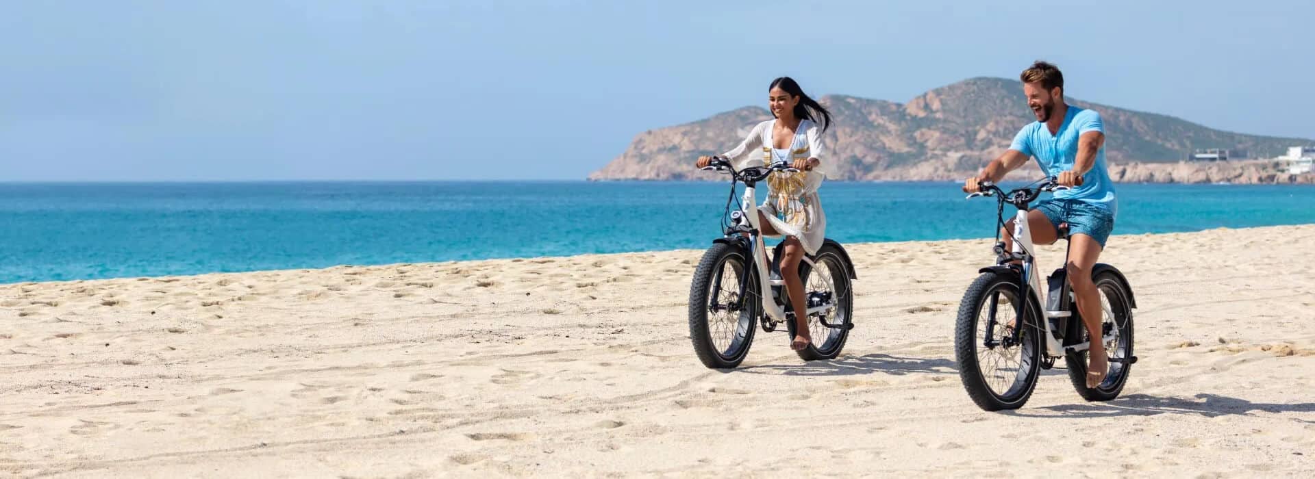a couple riding eBikes on the beach with the ocean and mountains in the background