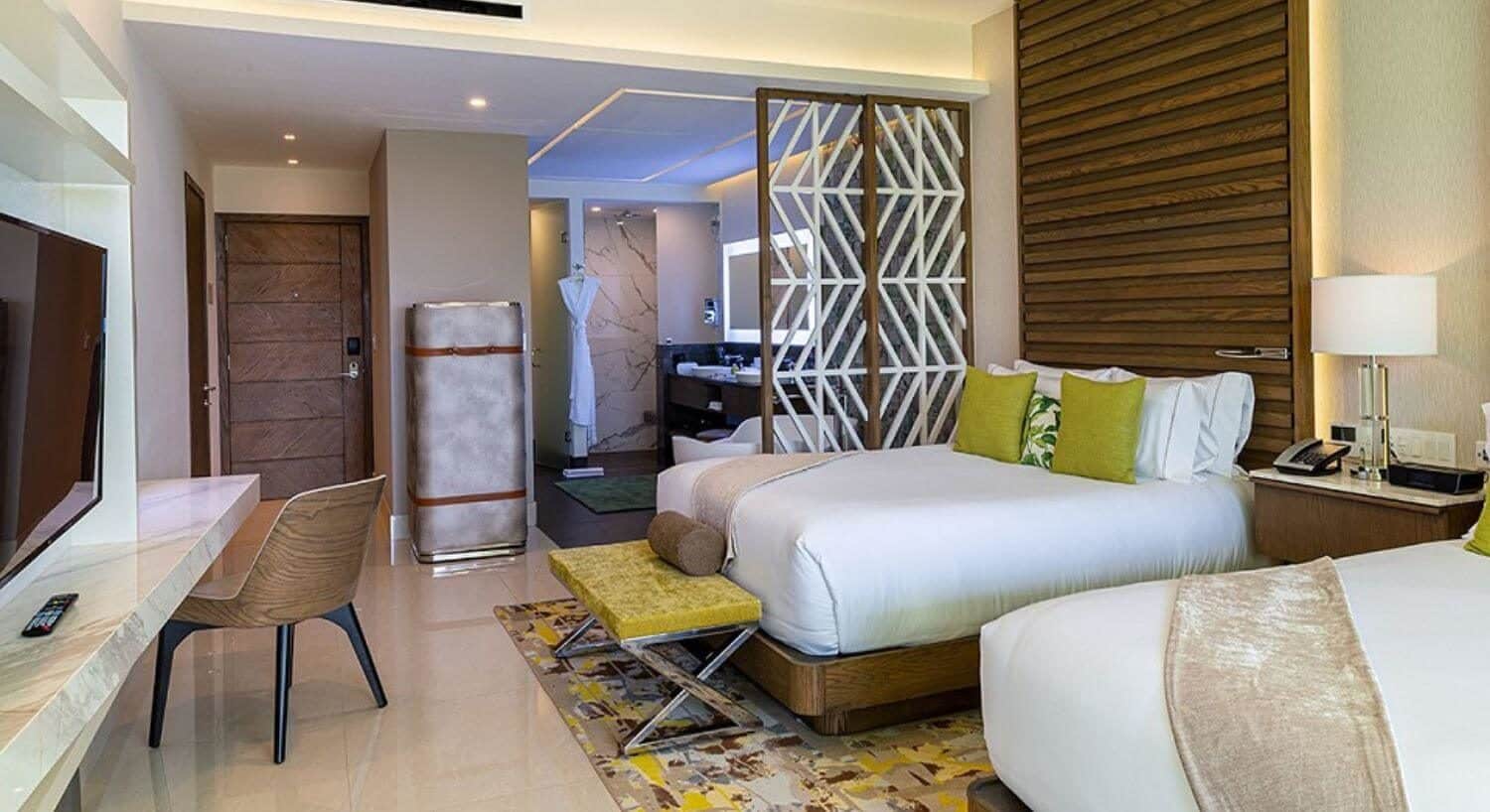 A bedroom with 2 queen beds, a flat screen TV on the opposite wall, a writing desk and chair with lit mirror above it, a tall stainless steel trunk on a wall near the beds, and a bathroom with deep soaking tub, walk in shower, and vanity with double sinks and lit mirror above the vanity.