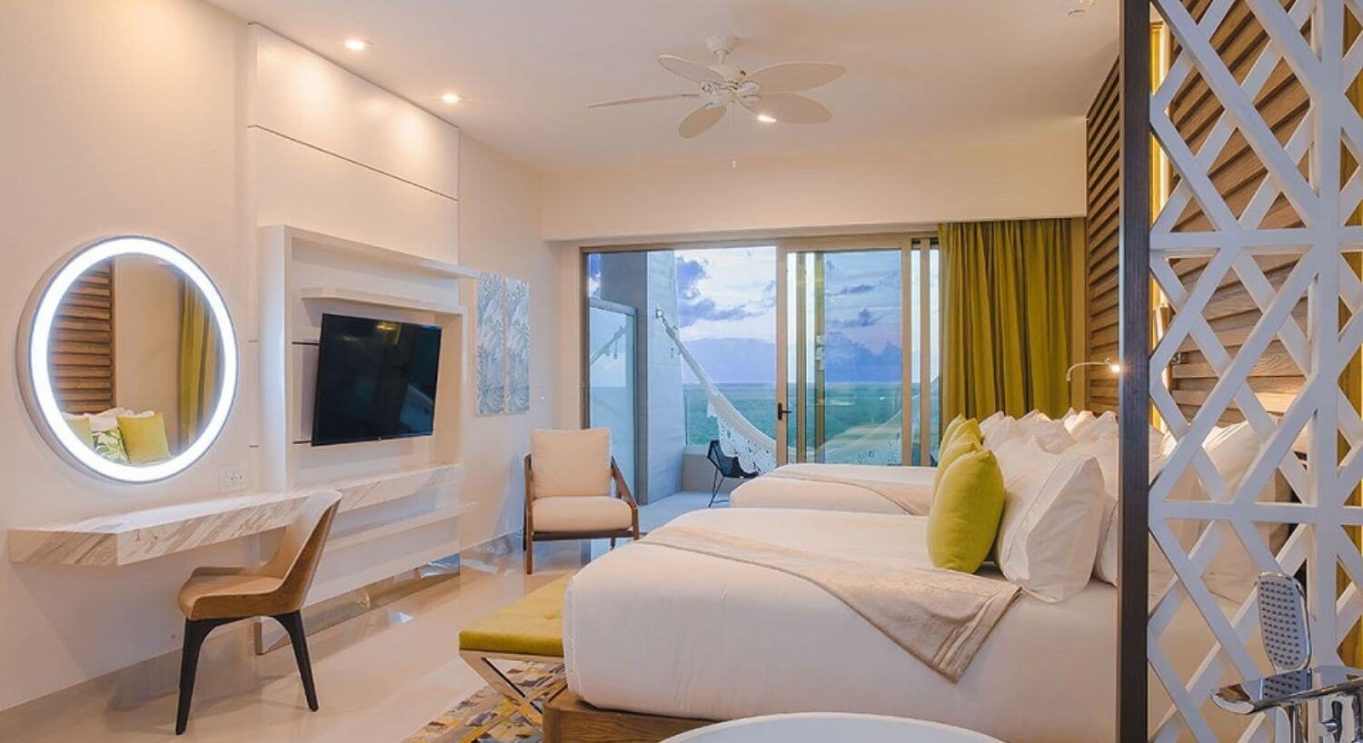 A bedroom with 2 queen beds, a sitting bench at the foot of one bed, a flat screen TV and writing desk with chair and mirror, a chair in the corner of the room, and sliding doors that lead out to a balcony with patio furniture and a hammock.