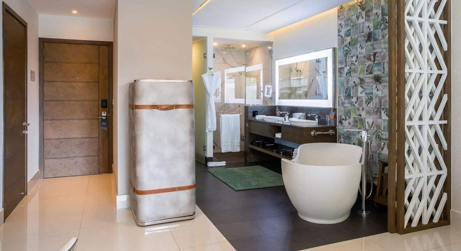 A room with a stainless steel trunk that opens to a hidden coffee center, and a bathroom with a deep soaking tub, a vanity with 2 sinks, and wide lit mirror above the vanity, and a walk in shower.