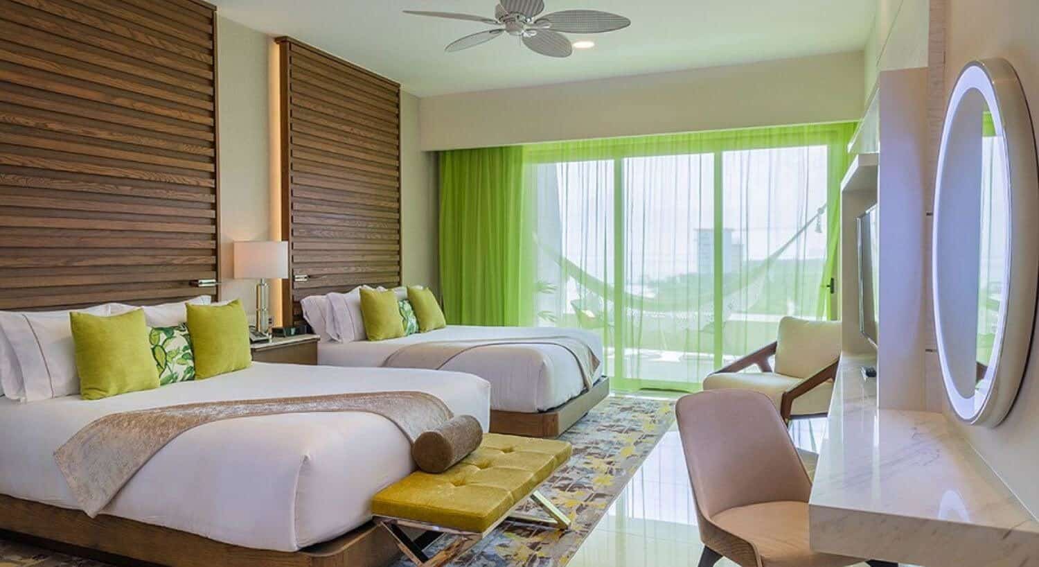 A bedroom with 2 queen beds with wood floor to ceiling headboards, a nightstand and lamp in between them, a sitting bench at the foot of one of the beds, a flat screen TvVand writing desk and chair on the opposite wall, and sheer green curtains over sliding glass doors that lead out to a balcony with patio furniture and a hammock.