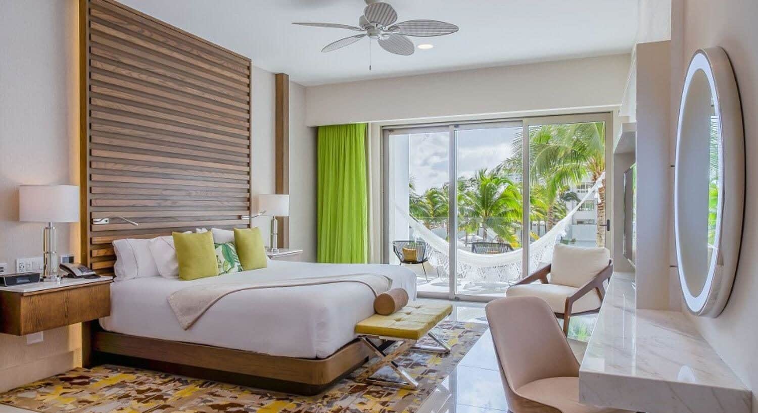 Bedroom with a bed with white bedding, a brown shiplap backboard, green and yellow pillows and curtains, a desk and chair, and sliding glass doors out to a patio with a hammock and the ocean in the background.