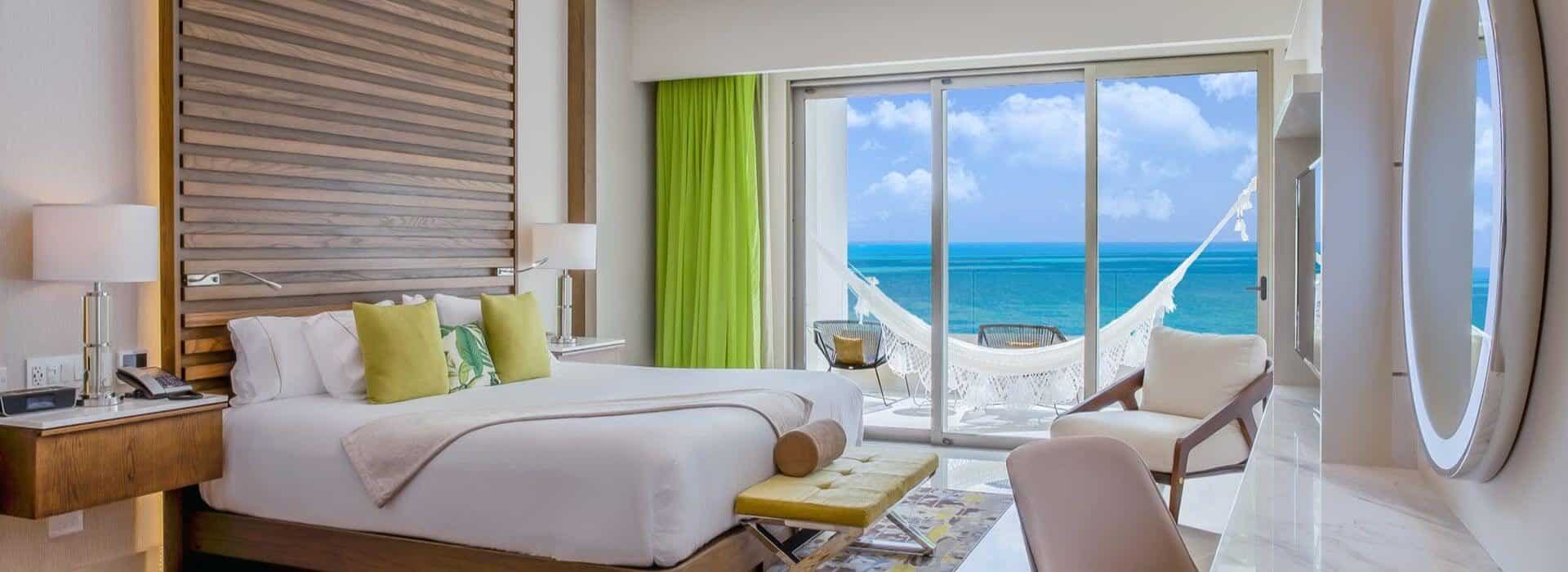 Bedroom with a king bed with white bedding and green accent pillows, nightstands on either side of the bed, a desk with chair and lighted mirror above the desk, and sliding glass doors opening to a patio with a hammock and ocean views.