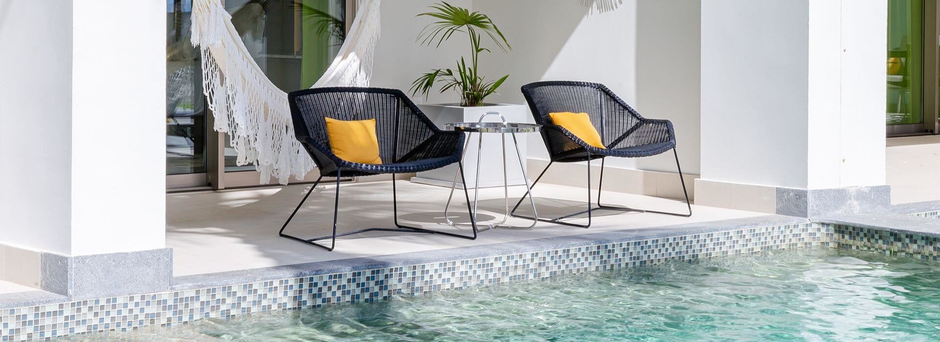 A patio with 2 wicker chairs with decorative pillows, a hammock, and a private swim up pool.