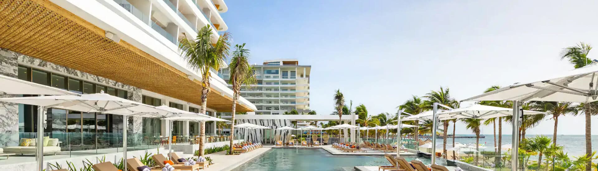 Outdoor pool at a hotel with lounge chairs, white umbrellas, palm trees, the ocean, and a multi-story building in the background.