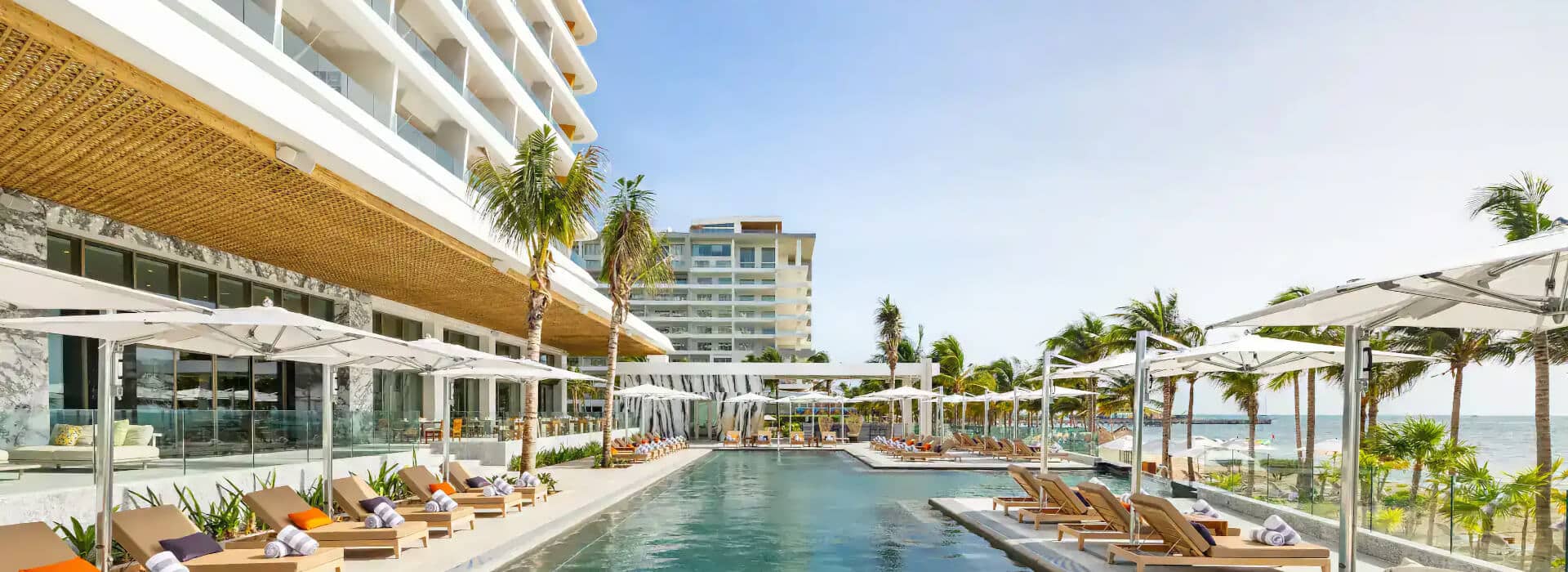 Outdoor pool at a hotel with lounge chairs, white umbrellas, palm trees, the ocean, and a multi-story building in the background.