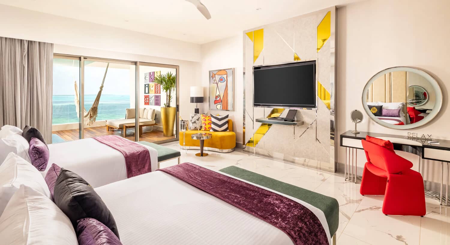 A hotel room featuring two beds with white linens and purple accents, a balcony with a hammock and ocean view, a television on a geometric wall, and a desk with the word 