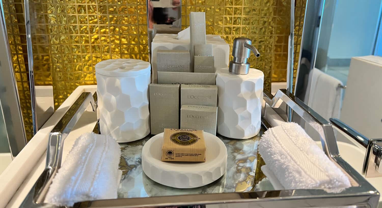 A silver tray holding various L'Occitane hotel bathroom amenities, including soaps, lotions, a soap dispenser, and folded towels, against a backdrop of a gold textured wall.