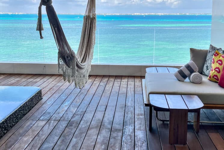 A balcony with wooden decking features a hammock, a cushioned bench with pillows, and a small table, all overlooking a bright turquoise ocean.