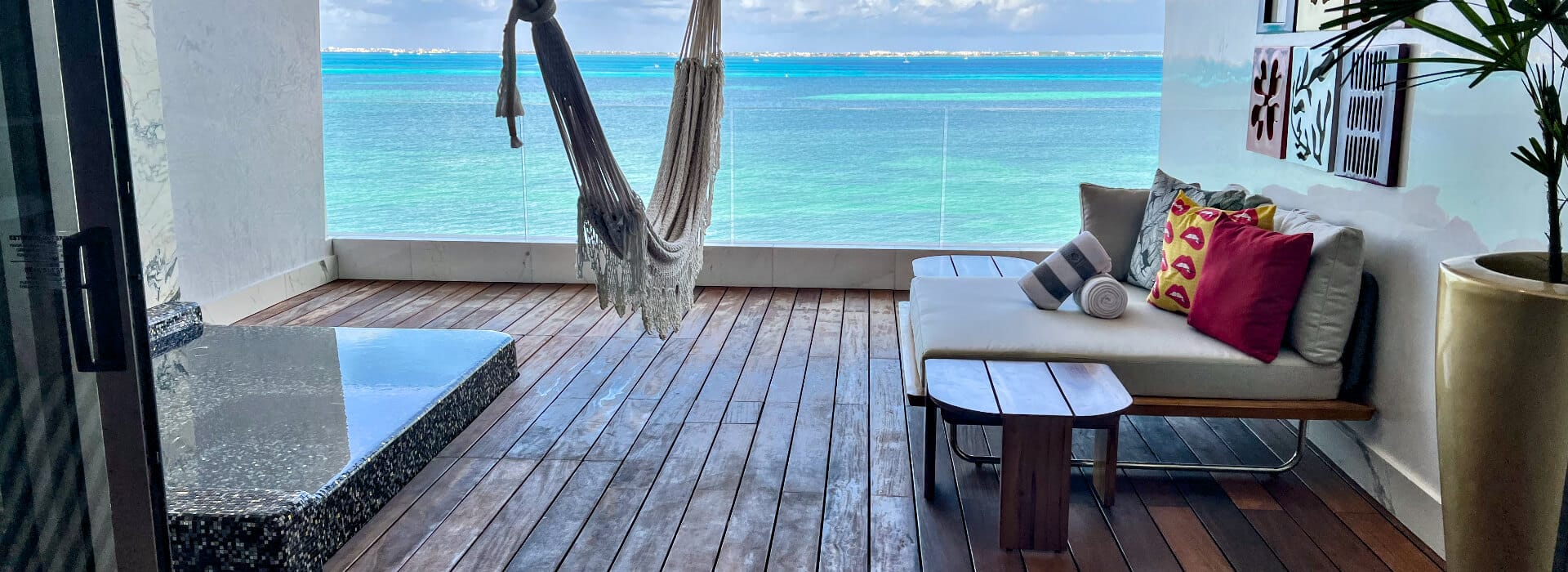 A balcony with wooden decking features a hammock, a cushioned bench with pillows, and a small table, all overlooking a bright turquoise ocean.