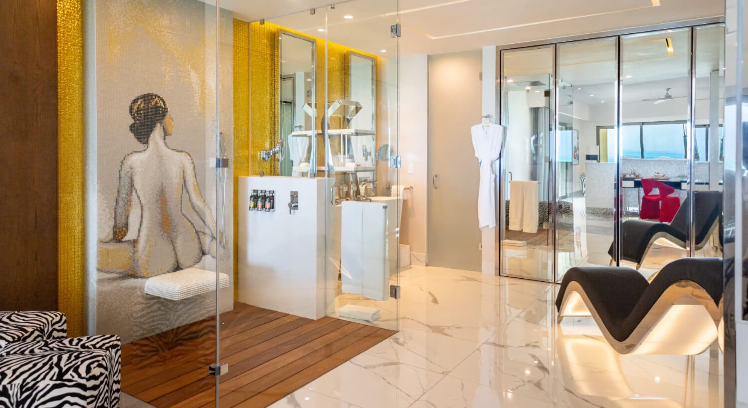 A hotel bathroom with a glass shower featuring a mosaic of a woman's back, a white robe hanging on a door, mirrored closet doors, and a view into a bedroom with a black tantra chair.