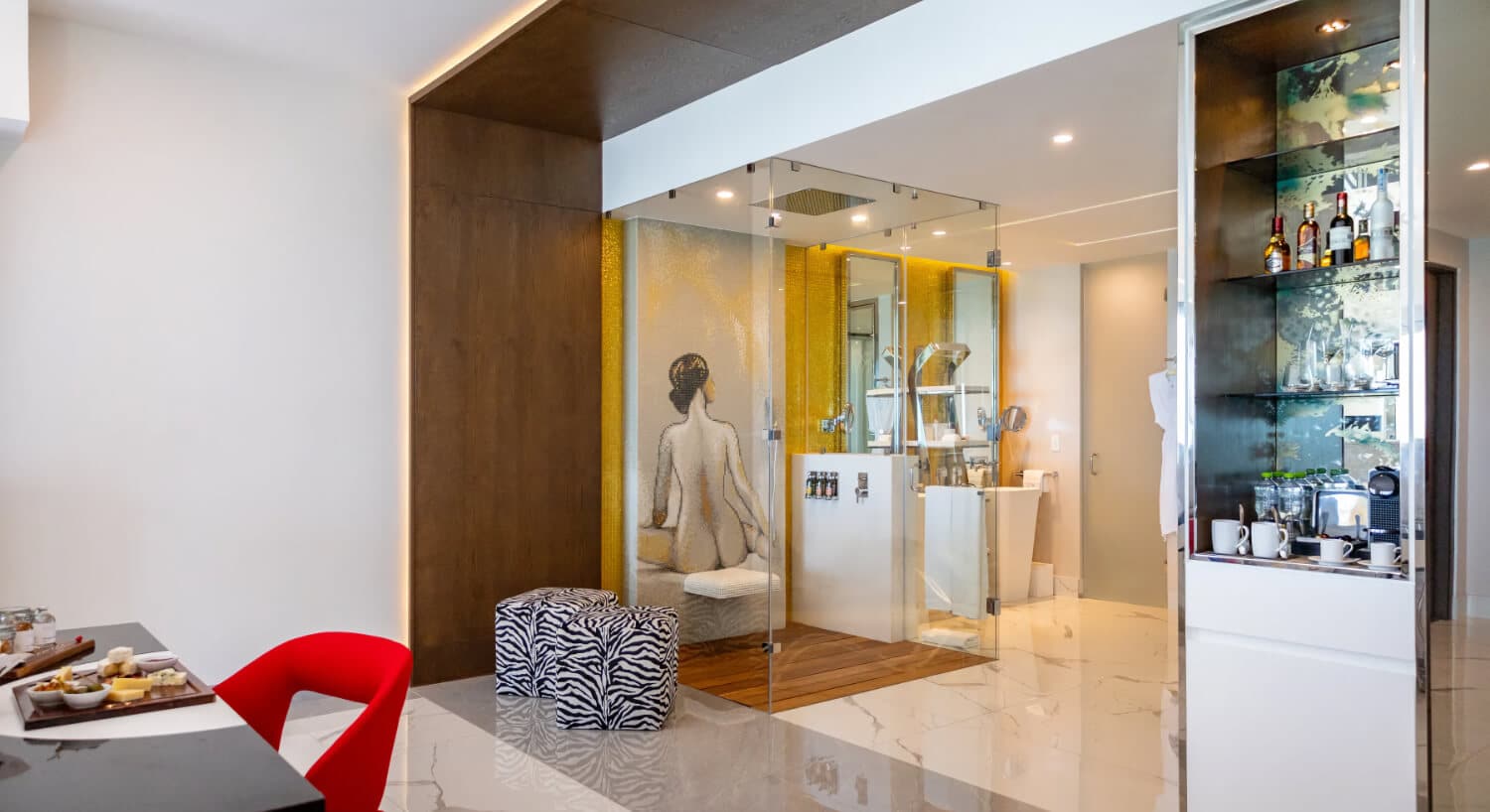 A hotel room with a bathroom featuring a glass shower with a mosaic of a woman's back, a yellow accent wall, a coffee center, and a desk with a charcuterie board and a red chair.