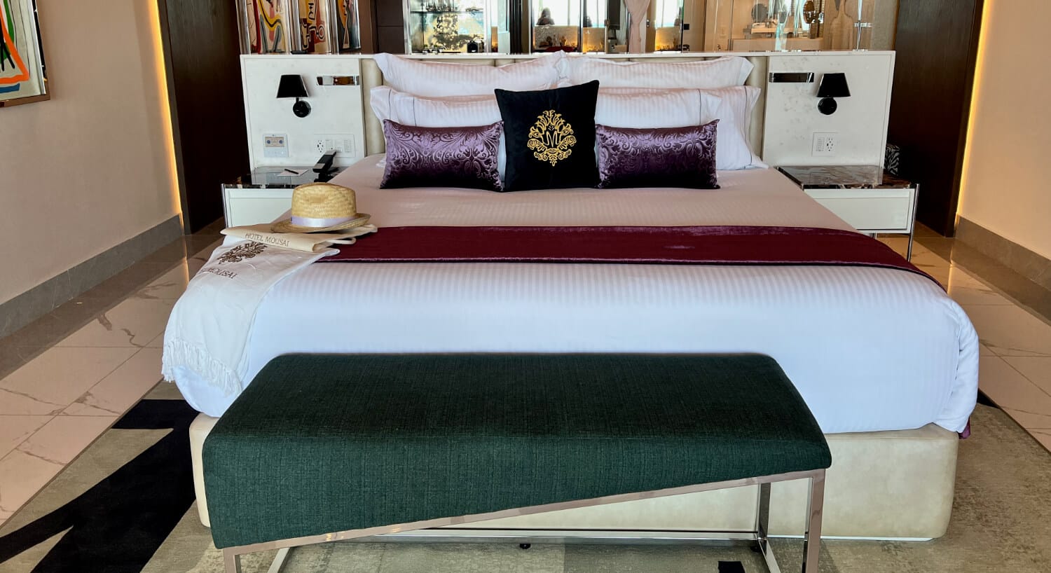 Luxury hotel room with a king bed with luxury linens and decorative pillows, a wrap and hat on the bed, and an upholstered bench at the foot of the bed.