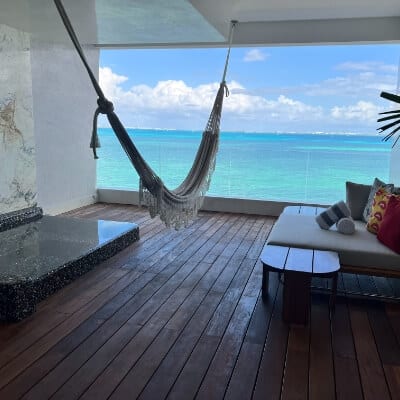 A modern balcony with dark wood decking and a white wall features a hanging hammock, a white cushioned bench with decorative pillows, a small wooden table, and a built-in jacuzzi, all facing a turquoise ocean.