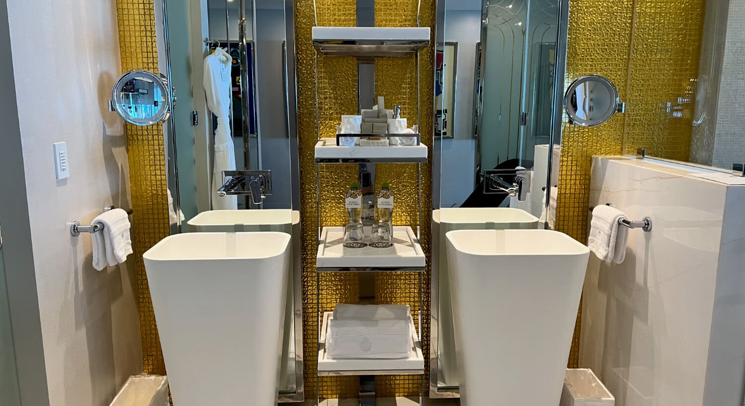 Luxury hotel bathroom with dual sinks, a gold accent wall, numerous shelves in between the sinks with L'Occitane amenities, water bottles, and towels, and a white bathrobe hanging nearby on a hook.