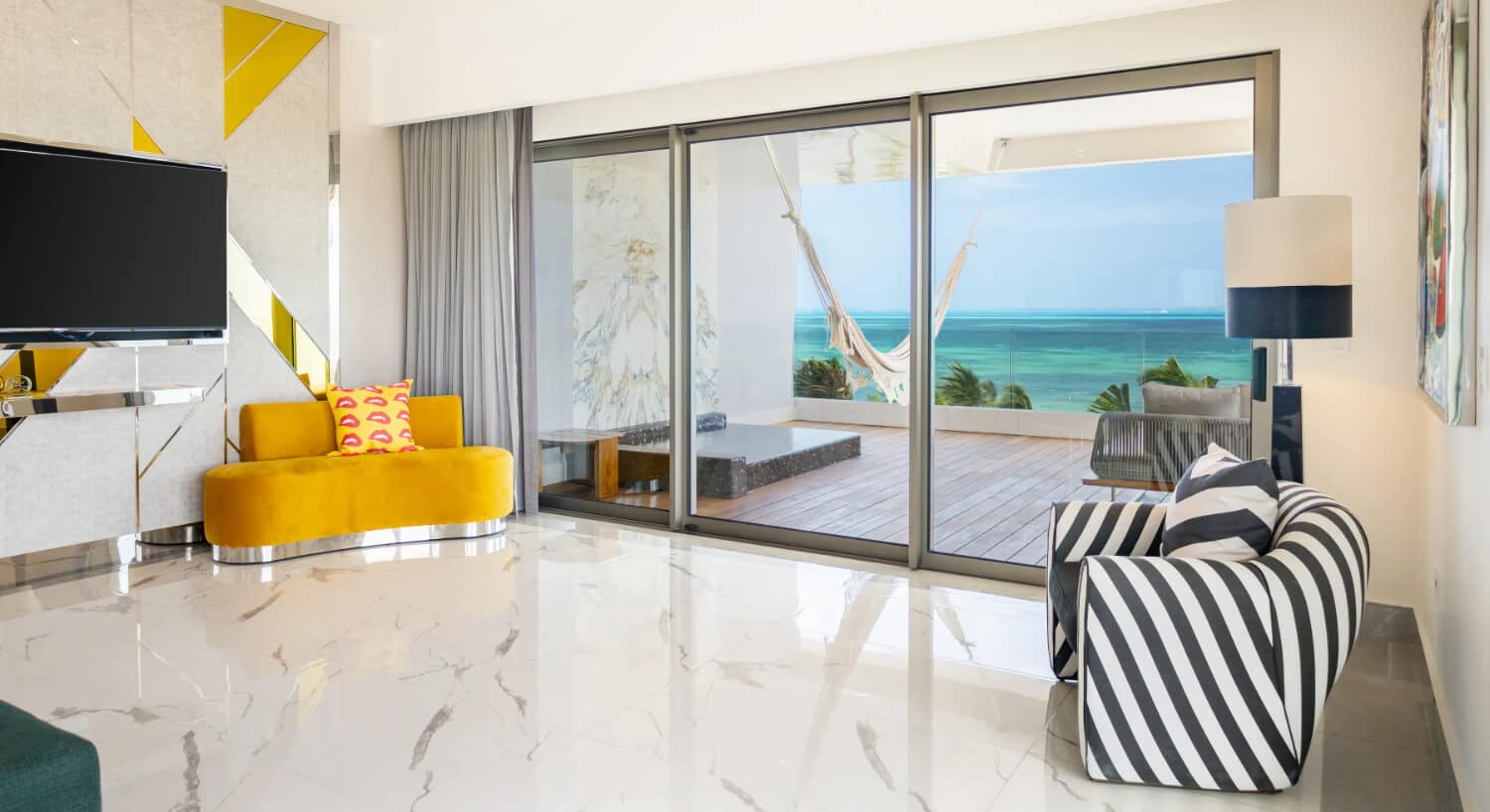 A hotel room with a yellow curved sofa, a television on a stand, a striped armchair, and a sliding glass door leading to a balcony with a hammock overlooking the ocean.