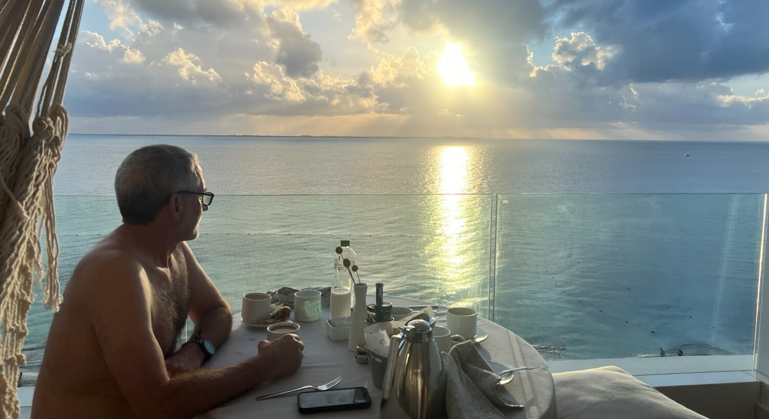 A man sitting at a table on a balcony overlooking a beautiful sunrise over the ocean while drinking coffee.