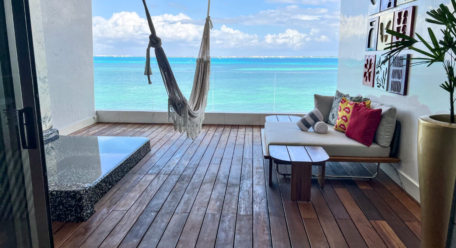 A modern balcony with dark wood decking and a white wall features a hanging hammock, a white cushioned bench with decorative pillows, a small wooden table, and a built-in jacuzzi, all facing a turquoise ocean.