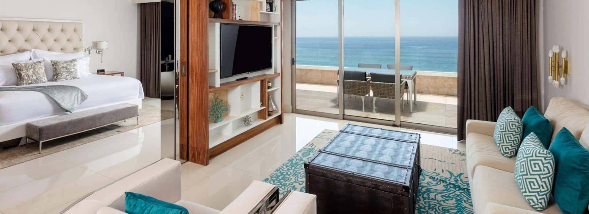 A living room and bedroom with sofa, tv, king bed, and sliding doors open to a patio with furniture overlooking the ocean