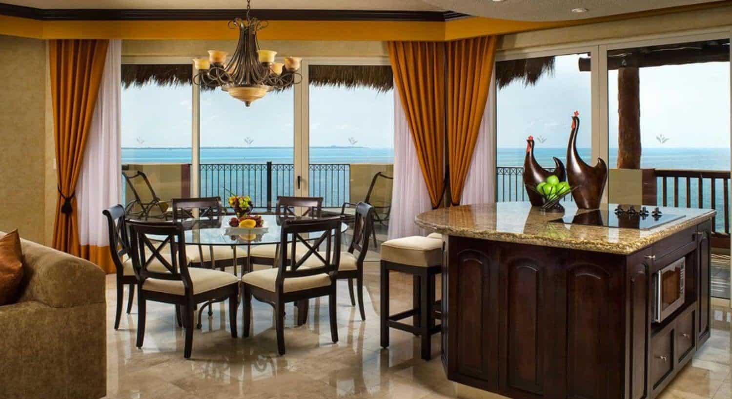 A spacious living area with a kitchen island, barstools, a round glass dining table with plush chairs, a sofa, and sliding doors that lead out to a wrap around terrace with sweeping ocean views.