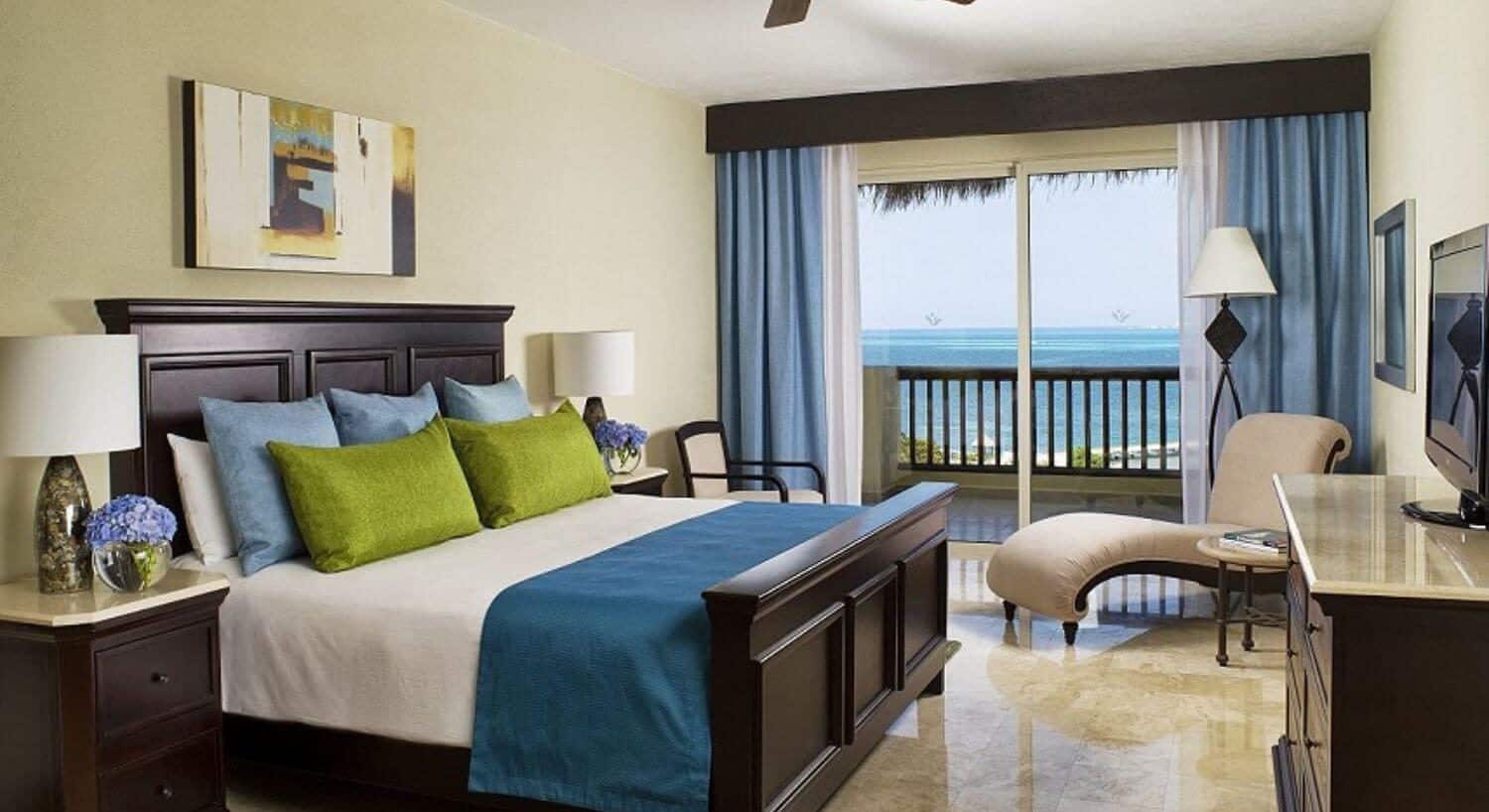 A bedroom with a King bed with white, glue and green bedding, a rich deep brown headboard and footboard, nightstands and lamps on either side of the bed, a dresser and flat screen TV on the opposite wall, a chaise lounge, and sliding doors that lead out to a balcony with ocean views.