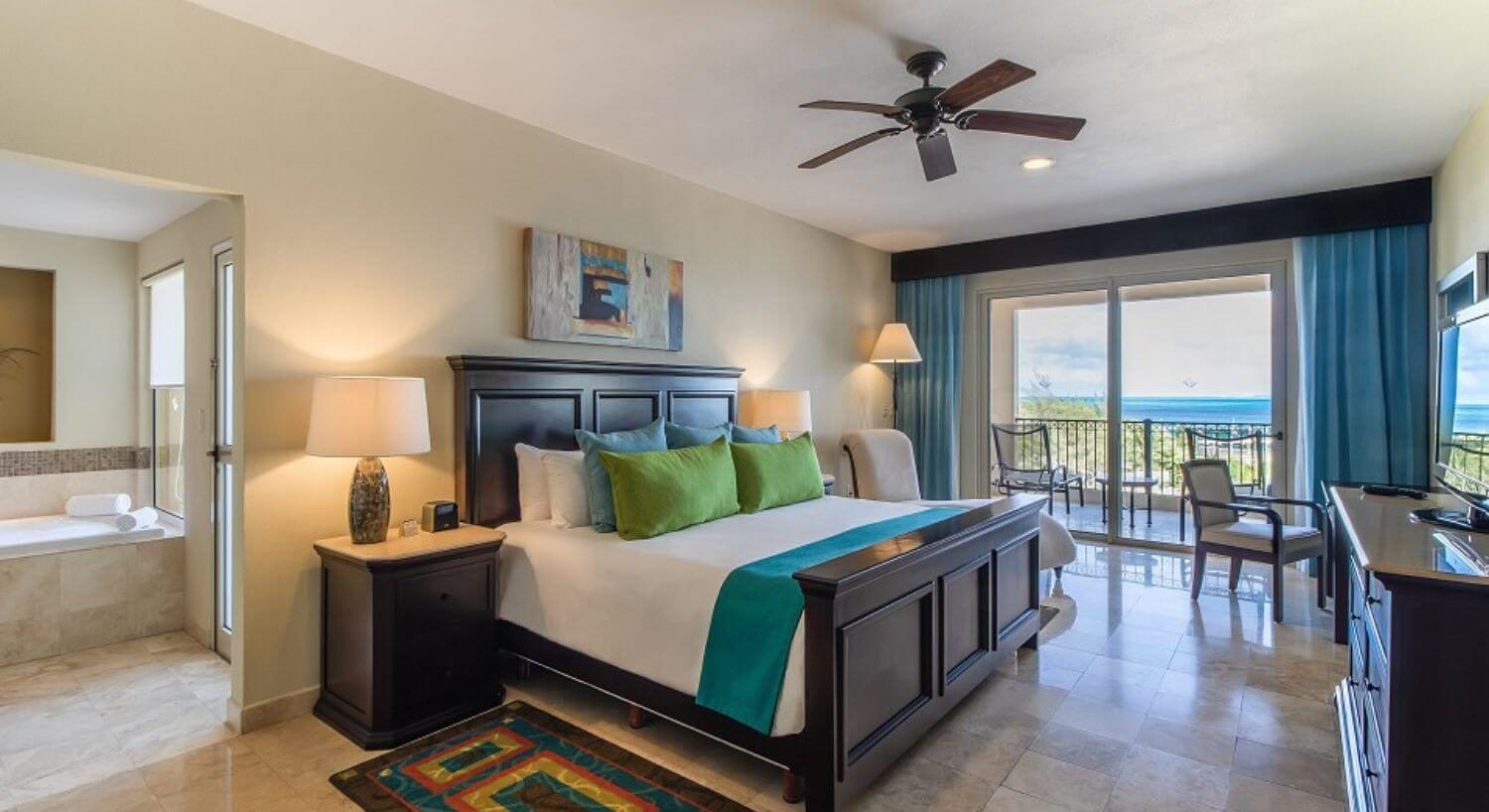 A bedroom with a King bed with dark wood headboard and footboard, white, green and blue bedding, matching nightstands and lamps on either side of the bed, a dresser with TV on the opposite wall, a desk and chair, sliding doors leading out to a balcony with patio furniture and ocean views, and a bathroom next to the bed with a jetted tub.