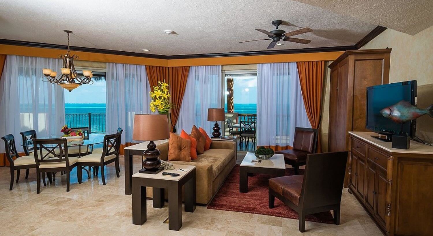 A comfortable living room with a plush sofa, two leather chairs, a marble coffee table and end tables, a hutch with a flat screen TV, a tall murphy bed, and dining area with a round glass table and several dining chairs. Sliding doors on two sides lead out to a wrap around terrace outdoor furniture and sweeping views of the turquoise Caribbean sea.