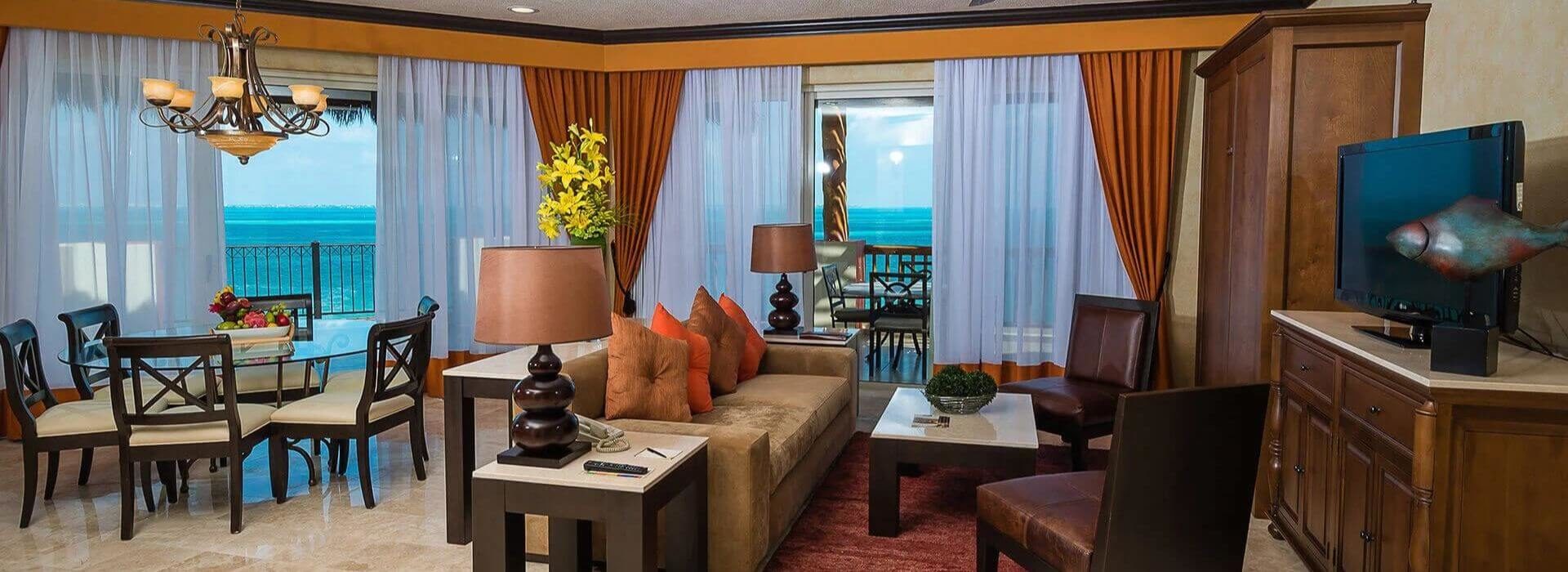 A comfortable living room with a plush sofa, two leather chairs, a marble coffee table and end tables, a hutch with a flat screen TV, a tall murphy bed, and dining area with a round glass table and several dining chairs. Sliding doors on two sides lead out to a wrap around terrace outdoor furniture and sweeping views of the turquoise Caribbean sea.