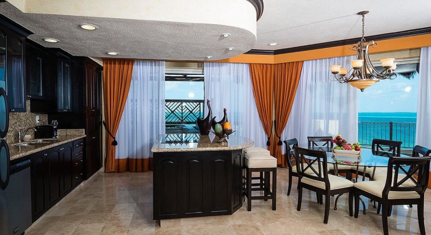 A gourmet kitchen and dining area with dark wood cupboards, granite countertops, an island with barstools, a round glass dining table with comfortable chairs, and sliding doors leading out to a wrap around terrace with a hot tub and the turquoise waters of the Caribbean sea.
