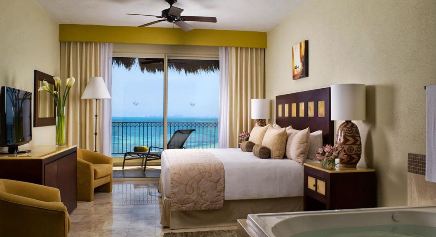 A bedroom with a King bed with white and tan bedding, a dark wood headboard, a dresser and flat screen TV on the opposite all, two comfortable armchairs, and part of a jacuzzi tub in the foreground. Sliding glass doors open out to a terrace with patio furniture and views of the the turquoise ocean.