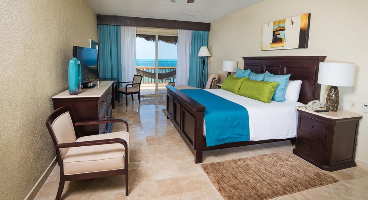 A bedroom with a King bed with white, blue and green bedding, deep wood headboard and footboard, nightstands with lamps, a dresser with TV an armchair, a desk and chair, and sliding doors that open to a terrace with hammock and ocean views.