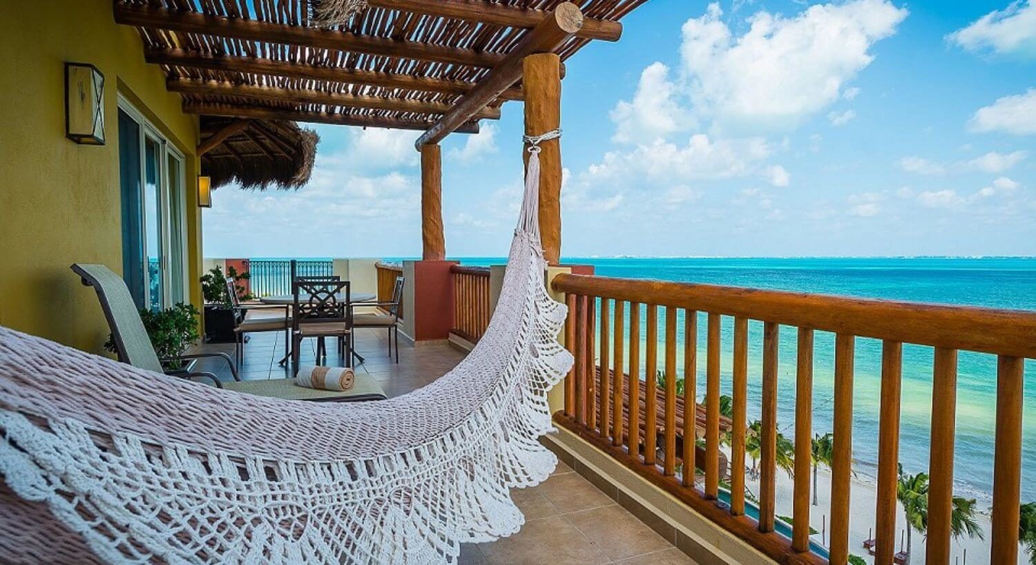 A long balcony with outdoor patio furniture, a hammock, lounge chairs, wood railings, and sweeping turquoise ocean views.