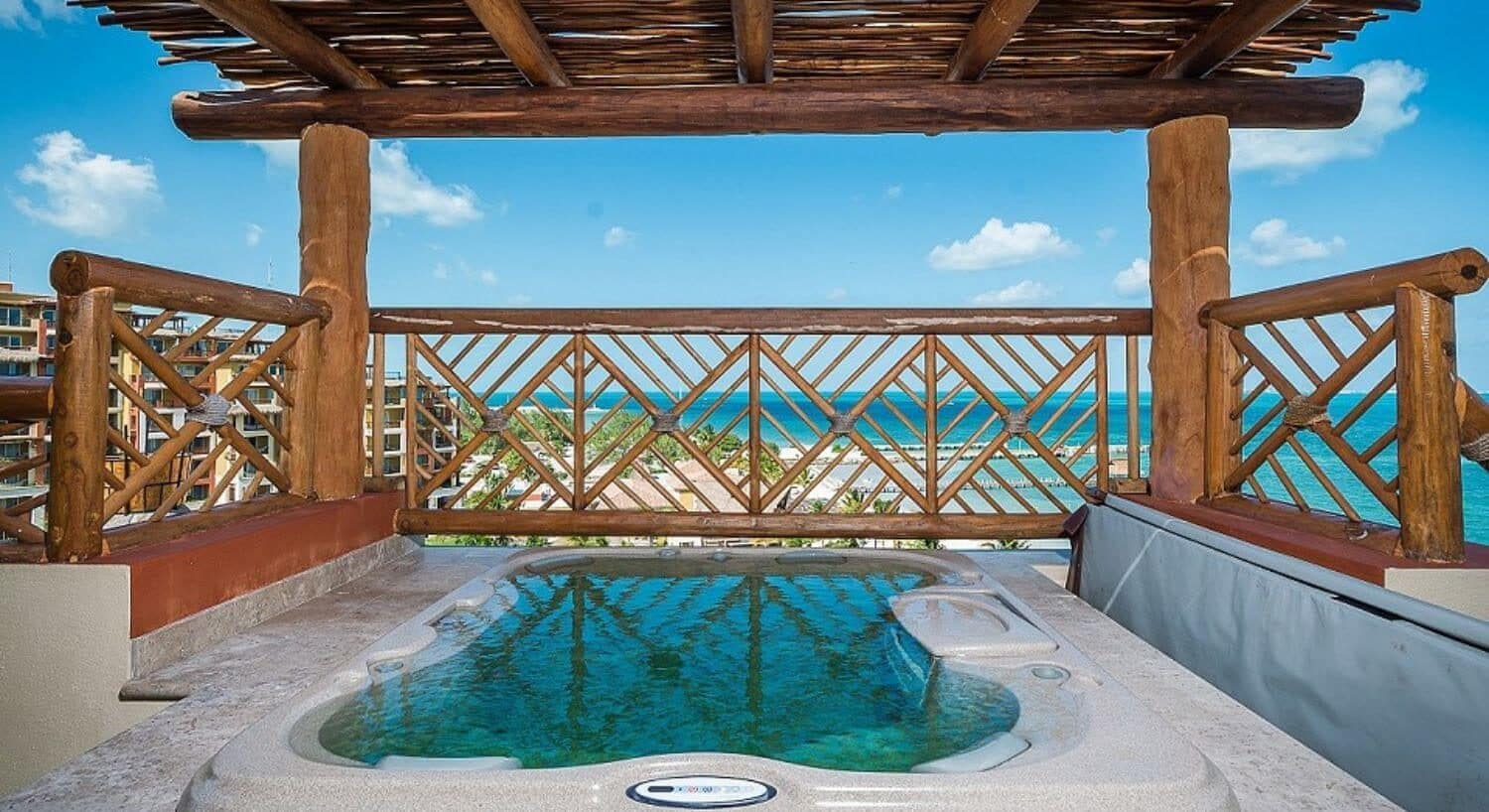 A crystal clear blue hot tub with a pergola roof and a wood railing around the hot tub with the tuquoise ocean in the background.