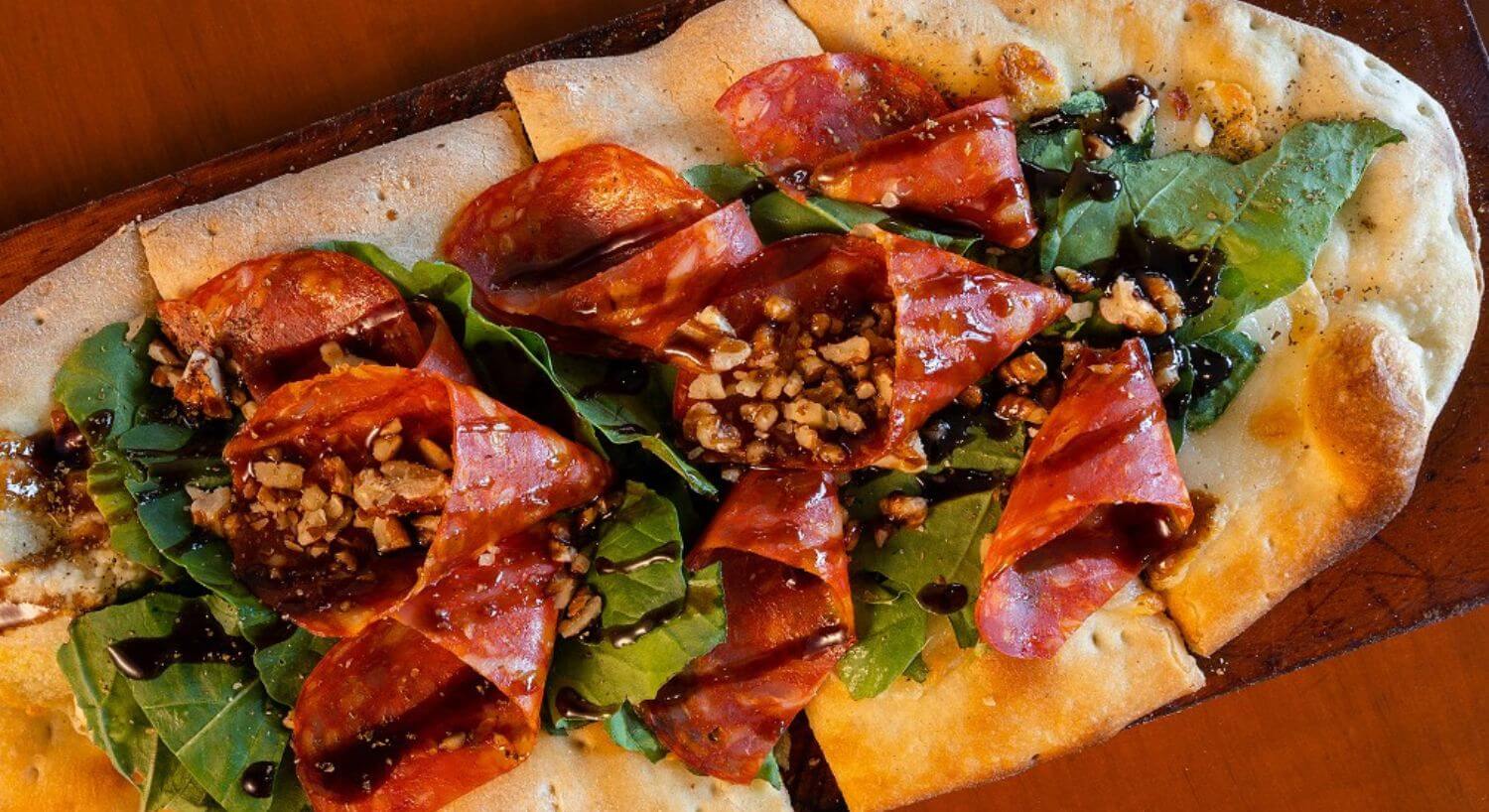 An oblong piece of flatbread cut into slices and topped with fresh basil leaves, slices of prosciutto, toasted njts, and drizzled with a balsamic glaze.