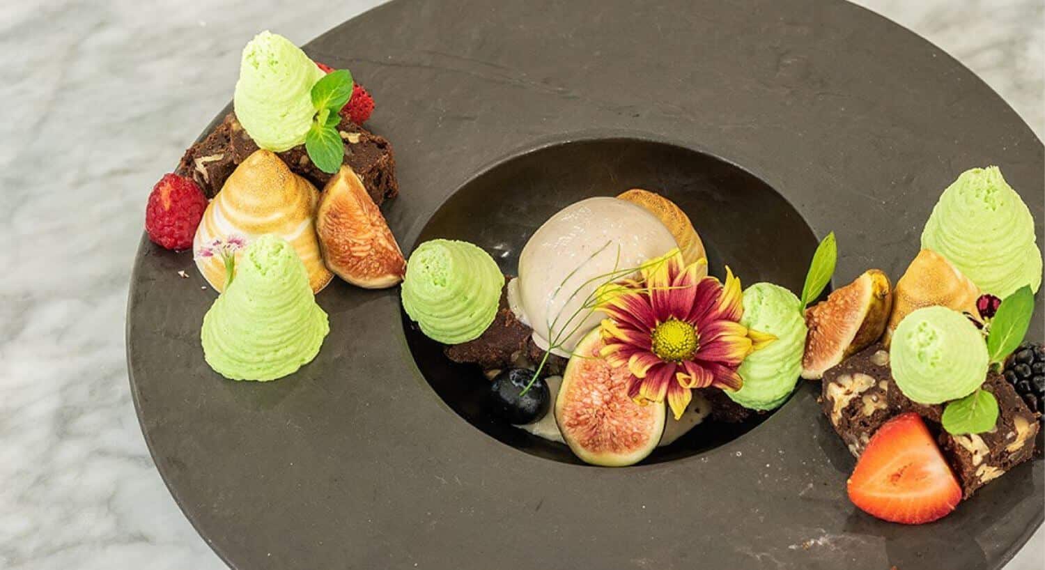 A grey plate and black bowl filled with ice cream and various fruits, brownies, and edible flowers.